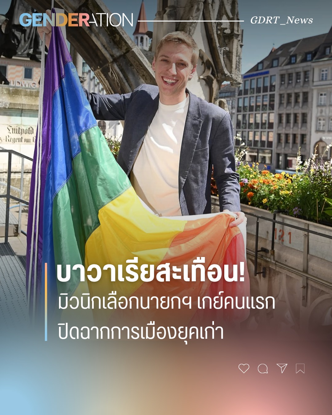 Young man in a blazer smiles while holding a rainbow Pride flag outdoors, with a city square and flowers in the background.